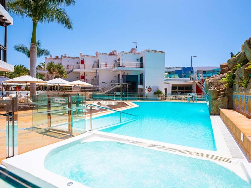 Modern hotelzwembad met ligstoelen, parasols en palmbomen onder een heldere lucht.