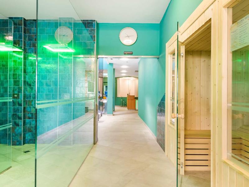 Heldere wellnessruimte met glazen douche en houten sauna in hotelgang met turquoise muren.
