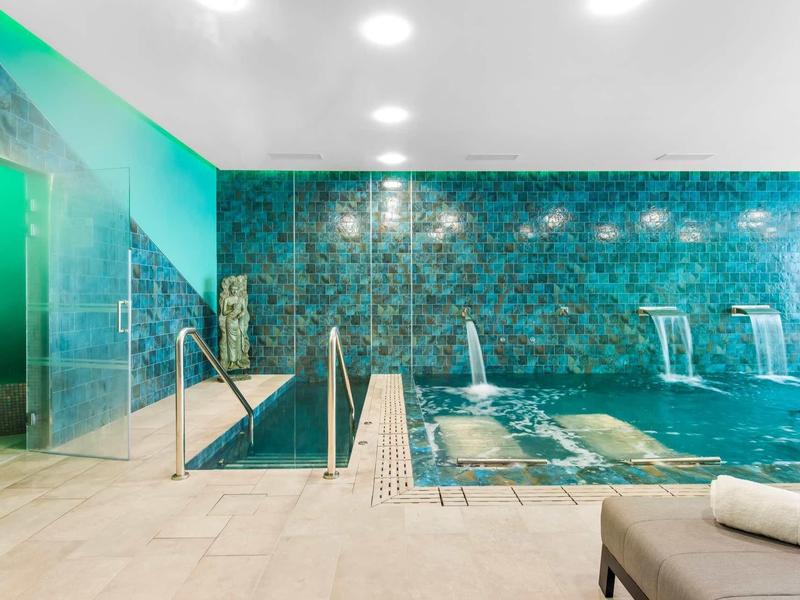 Binnenzwembad met turquoise tegels, waterstralen en ligstoelen in een moderne wellnessruimte.