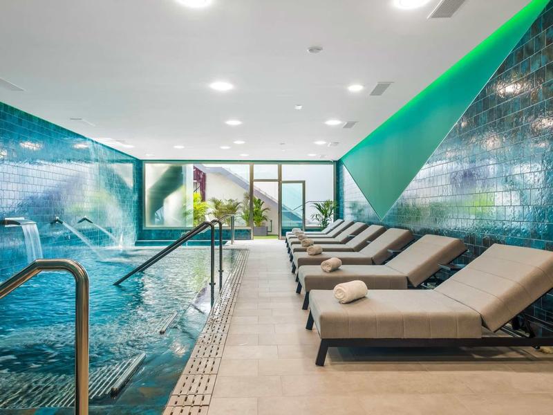 Binnenzwembad met meerdere watervallen en ligstoelen in een moderne wellnessruimte.