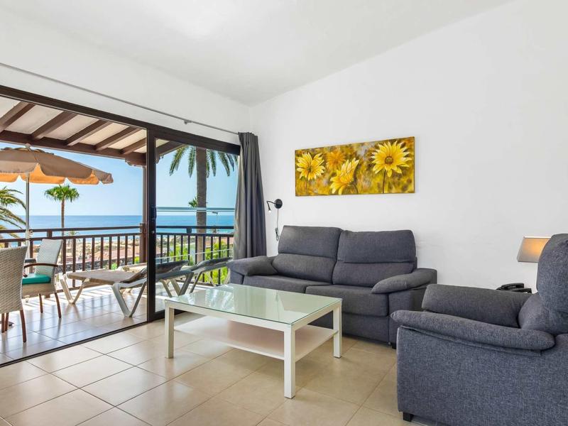 Lichte woonkamer met bank, fauteuil, glazen salontafel en balkon met uitzicht op zee.