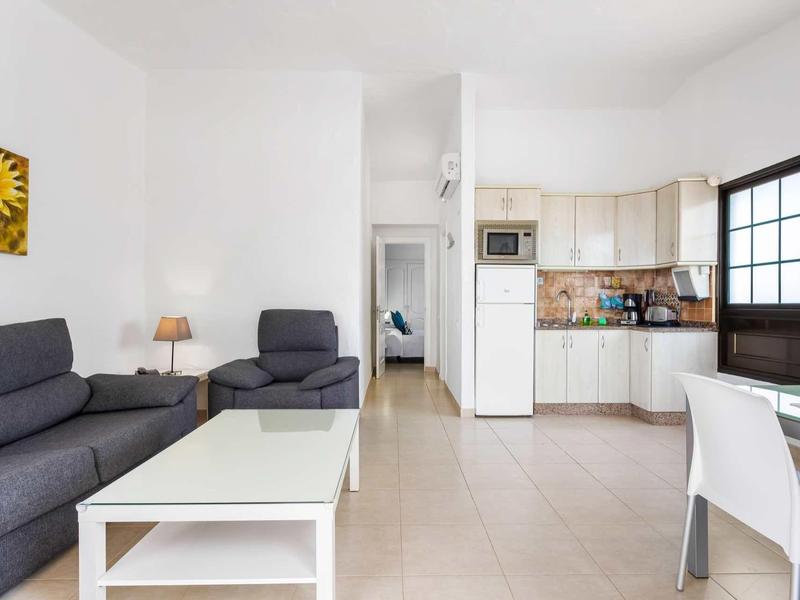 Woonkamer met grijze bank, fauteuil, witte tafel en geïntegreerde keuken in lichte ruimte