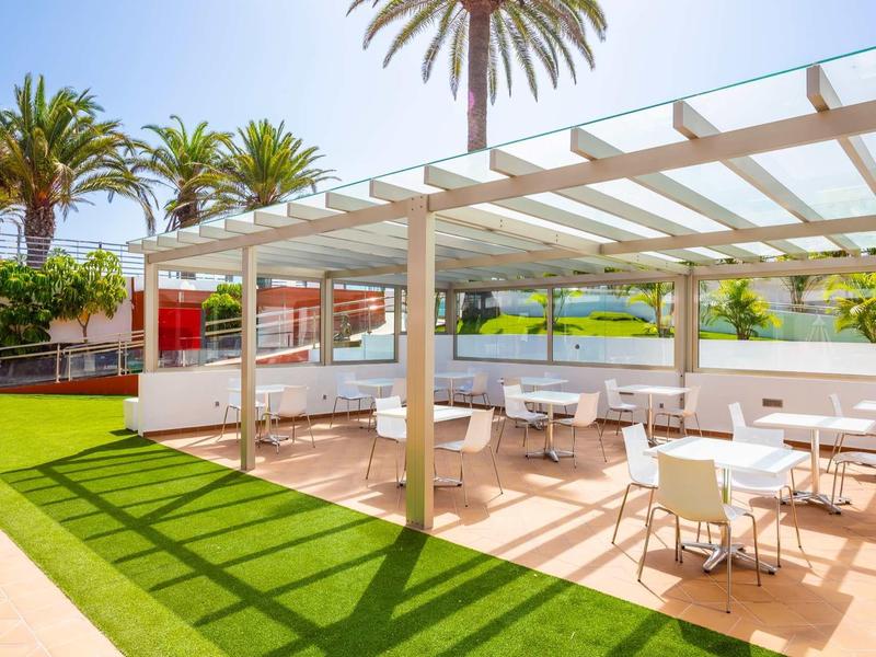 Buitenterras met witte tafels en stoelen naast groen gras onder een blauwe lucht.