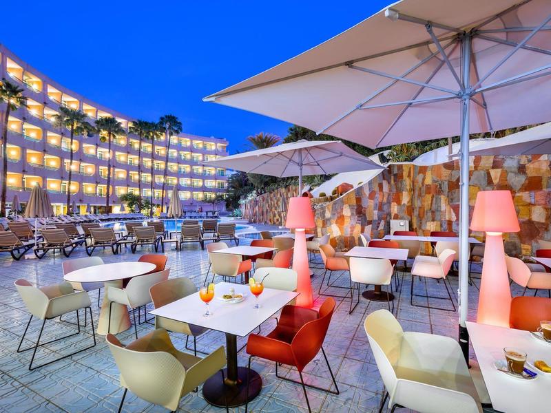 Hotelowy taras na zewnątrz z stolikami, krzesłami, parasolami i oświetlonym zakrzywionym budynkiem o zmierzchu.