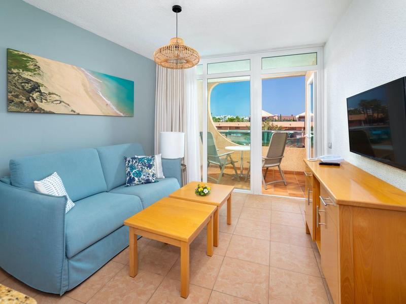 Przytulny salon z niebieską sofą, drewnianym stolikiem, telewizorem i balkonem z widokiem na morze.