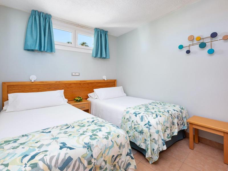 Pokój hotelowy z dwoma pojedynczymi łóżkami, kwiatowymi narzutami, drewnianym zagłówkiem, niebieskimi zasłonami i dekoracją ścienną.