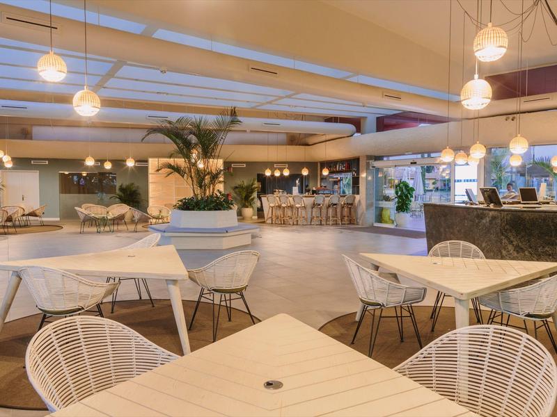 Area lounge d'hotel luminosa e moderna con tavoli, sedie e lampade a sospensione.