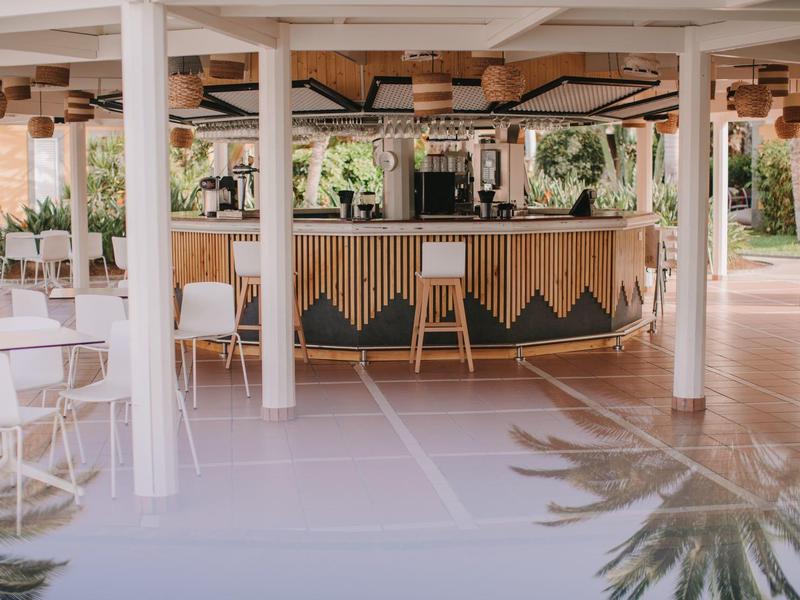 Bar all'aperto con bancone in legno e sedie bianche sotto un tetto aperto.