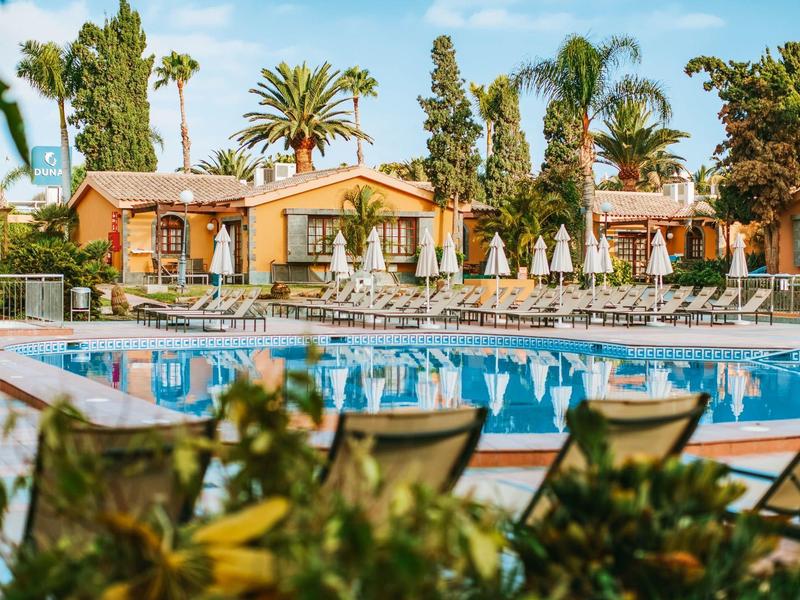 Piscina dell'hotel con lettini, ombrelloni e vegetazione tropicale sotto un cielo limpido.