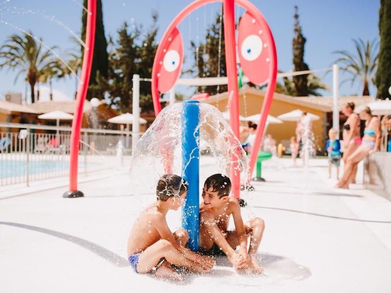Due bambini seduti vicino a un gioco d'acqua blu con figure rosse in un parco acquatico.