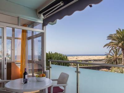 Table de balcon avec chaises donnant sur des palmiers et des dunes de sable sous un ciel clair.