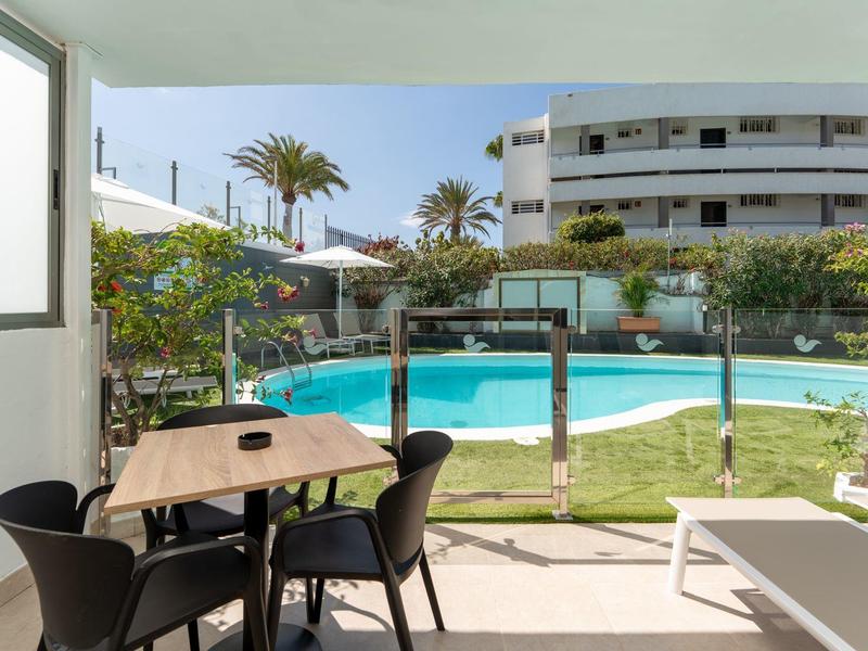 Blick von einer überdachten Terrasse mit Tisch und Stühlen auf einen Pool, umgeben von Palmen und Hotelgebäude.