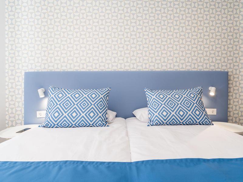 Modern double bed with blue pillows and headboard in a bright hotel room.