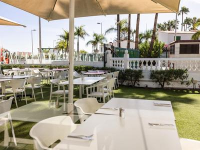 Área exterior soleada con mesas blancas, sillas y sombrillas sobre césped verde.