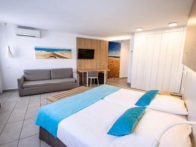 Habitación de hotel luminosa con cama doble, sofá, televisor y decoración moderna en tonos neutros.