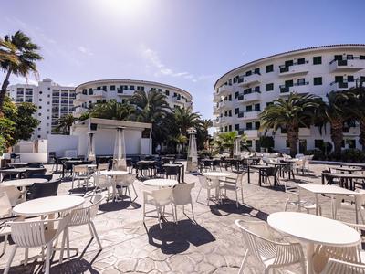 Terraza al aire libre con mesas y sillas blancas frente a edificios de hotel curvos bajo un cielo azul.
