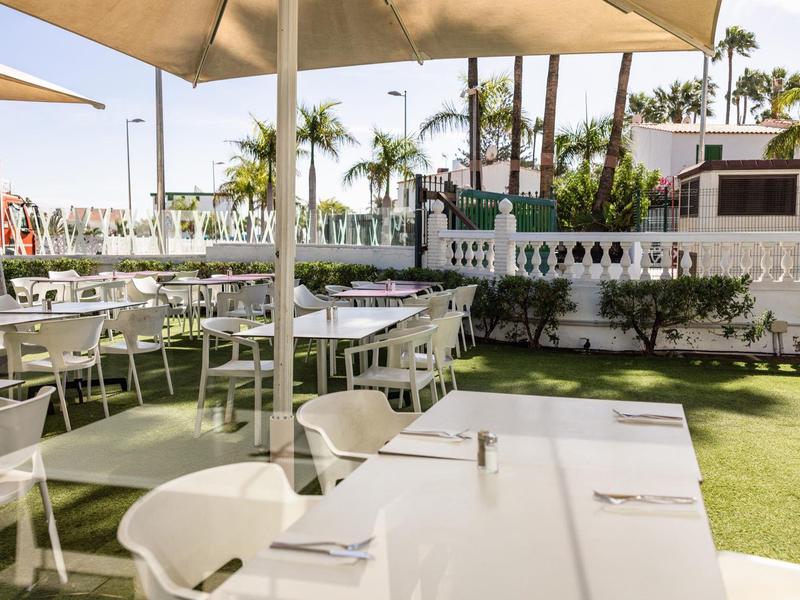 Área exterior soleada con mesas blancas, sillas y sombrillas sobre césped verde.