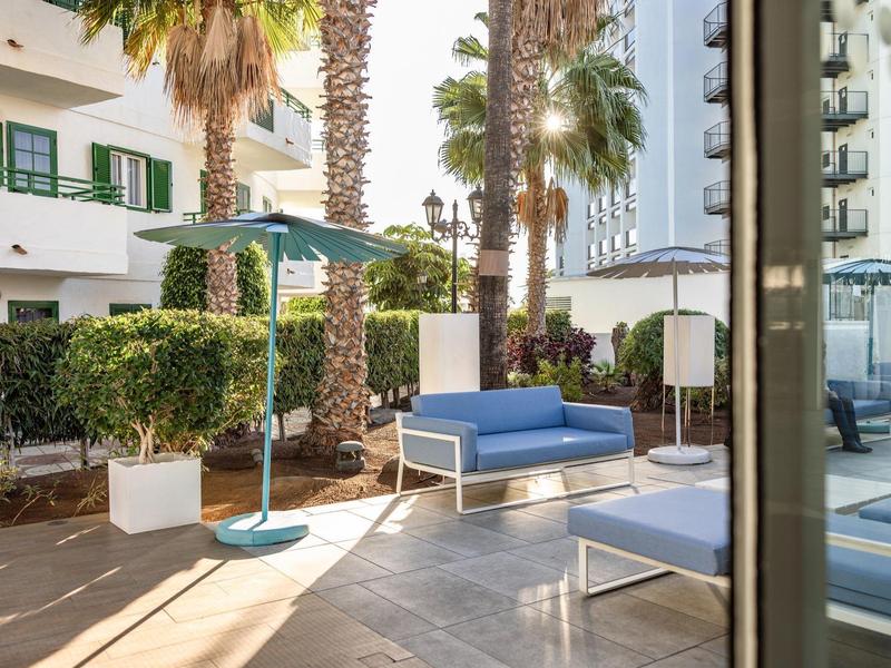 Área de terraza con sofás azules, sombrillas y palmeras junto a un edificio blanco con ventanas verdes.