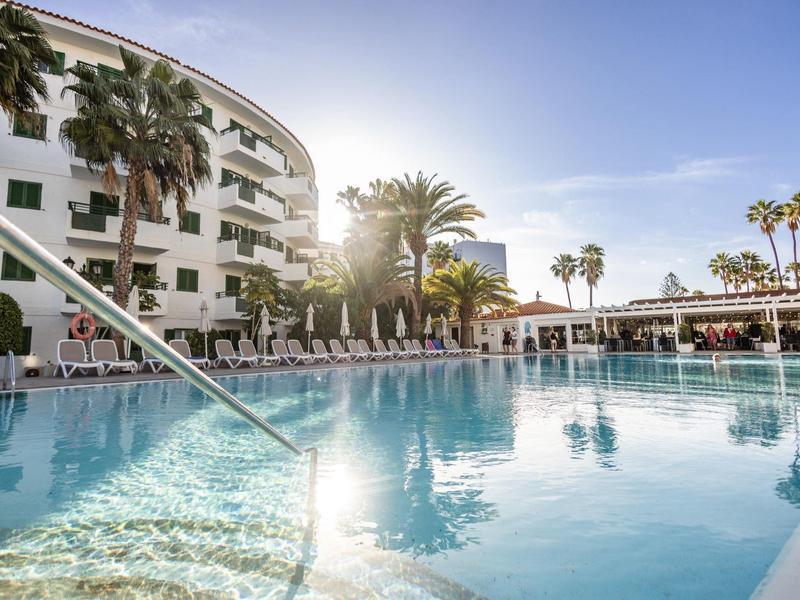 Gran piscina con tumbonas y palmeras en un resort hotelero con clima soleado.
