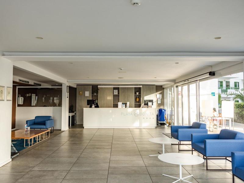 Luminosa recepción de hotel con mostrador, sillones azules y grandes ventanas con vista a la ciudad.