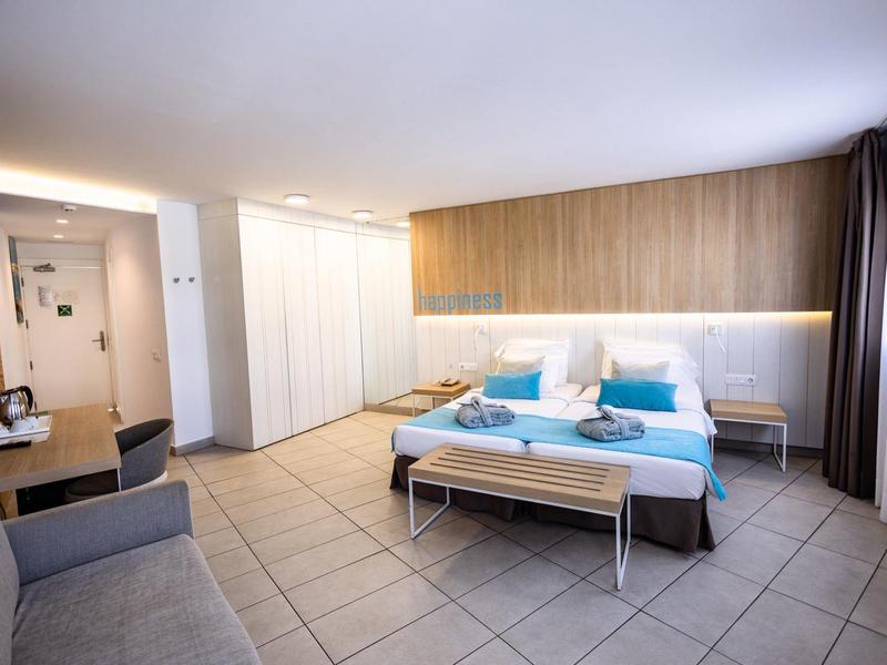 Habitación de hotel moderna con cama doble, sofá, armario y ventana grande con cortinas.