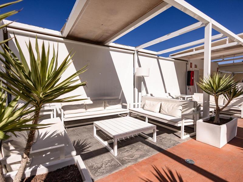 Área exterior moderna con muebles de salón blancos y plantas bajo un cielo azul.