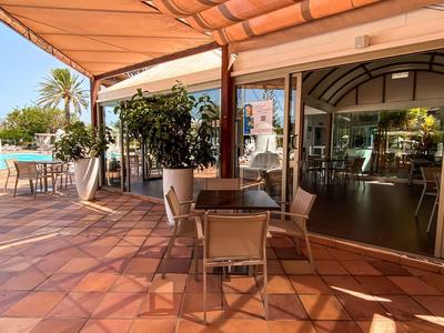 Terrasse avec sol carrelé beige, tables et chaises sous un auvent près d'une piscine.