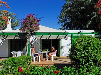 Zwei Personen sitzen an einem Tisch auf einer Terrasse vor einem weißen Haus mit grünem Dach bei sonnigem Wetter.