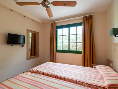 Chambre d'hôtel lumineuse avec deux lits rayés, ventilateur de plafond en bois et grande fenêtre avec rideaux.
