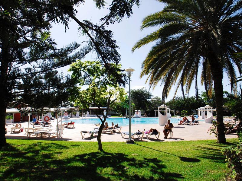Piscine extérieure ensoleillée entourée d'arbres, palmiers et de baigneurs relaxant sur des chaises longues