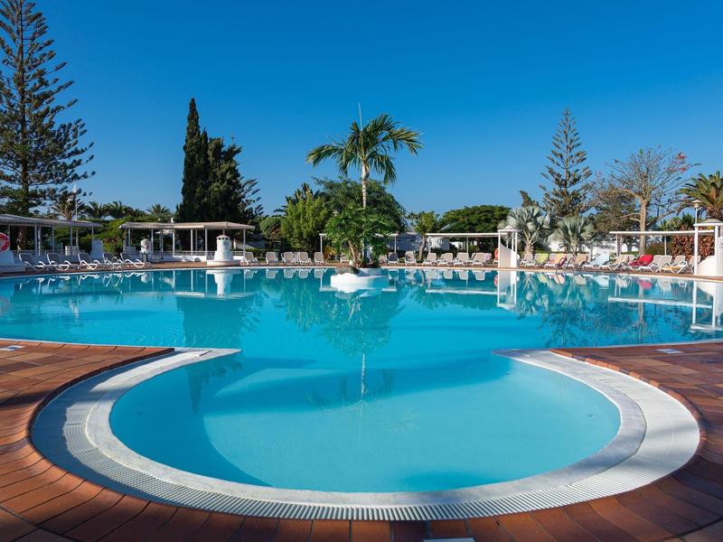 Grand piscine extérieure entourée de chaises longues et de palmiers sous un ciel bleu clair.