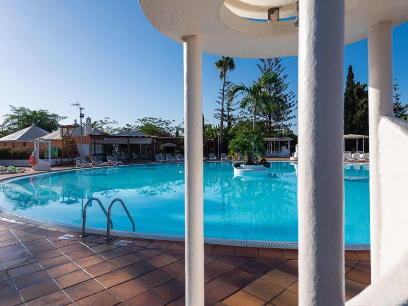 Piscine bleue claire entourée de palmiers et chaises longues sous un ciel ensoleillé.