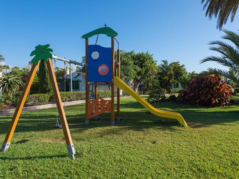 Aire de jeux avec balançoires, toboggan et palmiers par une journée ensoleillée dans un jardin d'hôtel.