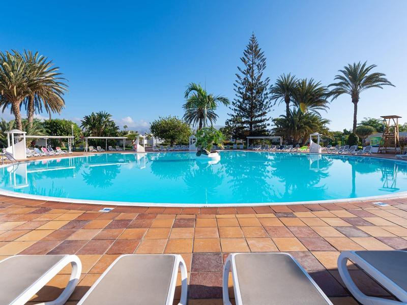 Piscine extérieure ronde avec eau claire, entourée de palmiers et de chaises longues sous un ciel dégagé.