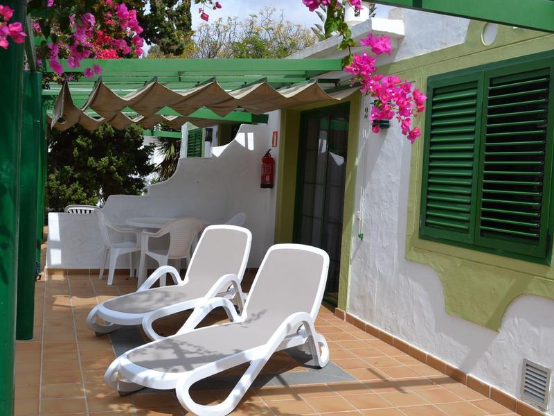 Terrasse ensoleillée avec transats blancs, volets verts et fleurs roses vives dans un cadre lumineux.
