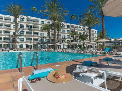 Piscine d'hôtel avec transats, parasols et palmiers sous un ciel bleu clair.