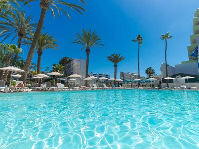 Klare blaue Poollandschaft mit Sonnenschirmen und Palmen im Hotelumfeld bei sonnigem Wetter.