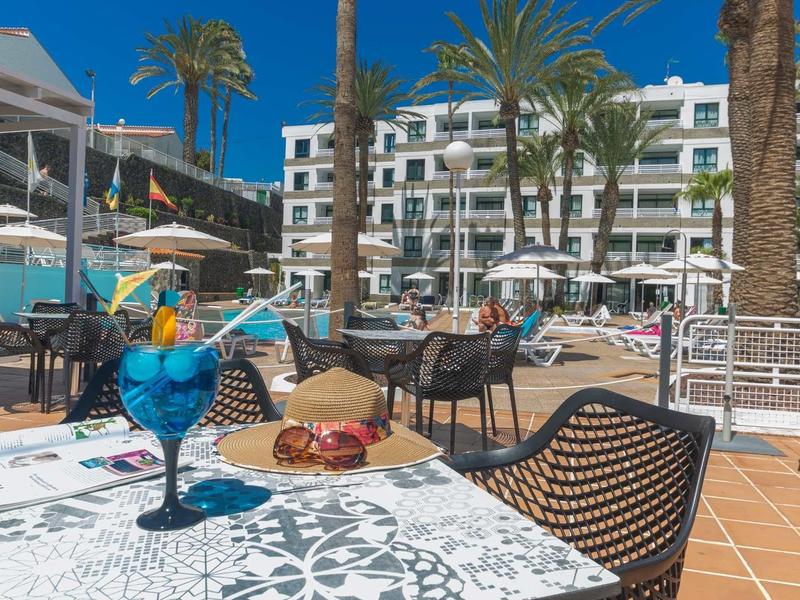 Terrasse avec tables, chaises et un cocktail devant un bâtiment hôtelier sous un ciel bleu.
