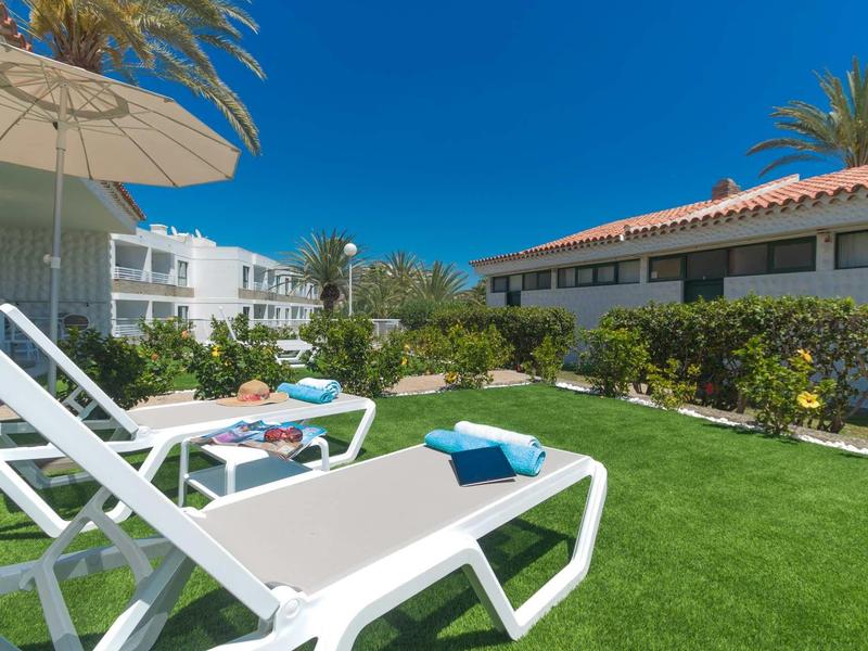 Chaises longues blanches avec serviettes bleues sur pelouse verte devant les bâtiments de l'hôtel par temps ensoleillé.