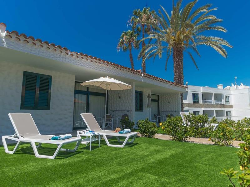 Chaises longues sur une pelouse entretenue devant une maison de vacances blanche avec des palmiers en arrière-plan.