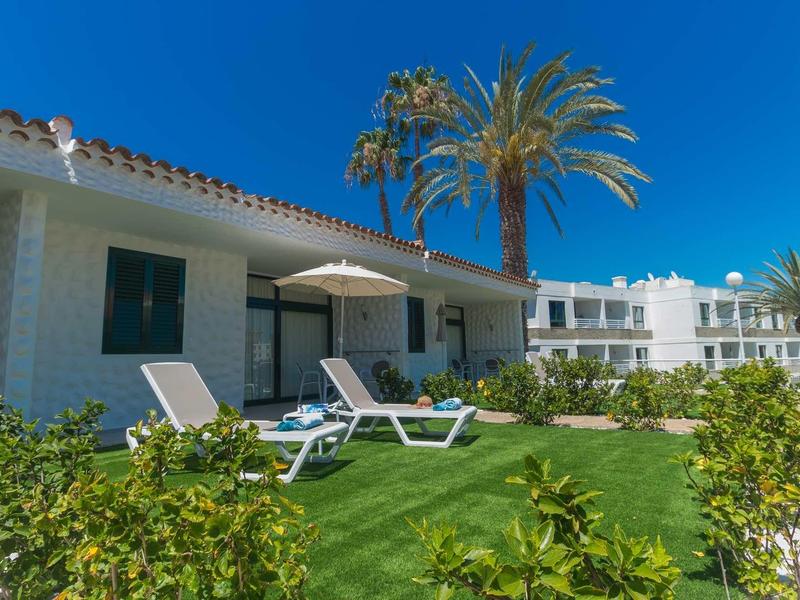 Terrasse ensoleillée avec chaises longues devant une maison de vacances blanche et des palmiers.