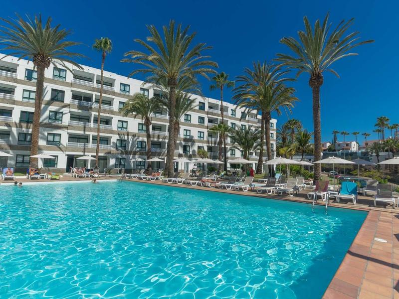 Grand piscine extérieure devant un hôtel à plusieurs étages sous un ciel bleu avec des palmiers et des chaises longues.