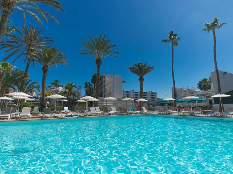 Grande piscine à l'eau claire, entourée de chaises longues et de palmiers sous un ciel bleu.