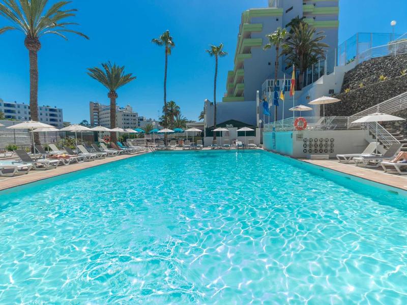 Grande piscine extérieure avec chaises longues et palmiers sous un ciel bleu clair dans un hôtel.