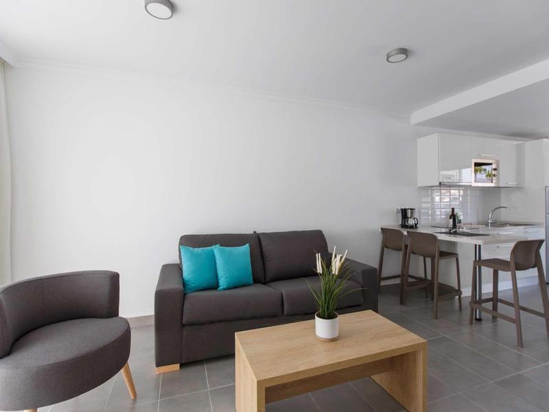 Salon moderne avec canapé gris, coussins bleu-vert, fauteuil et table en bois près du bar de cuisine.