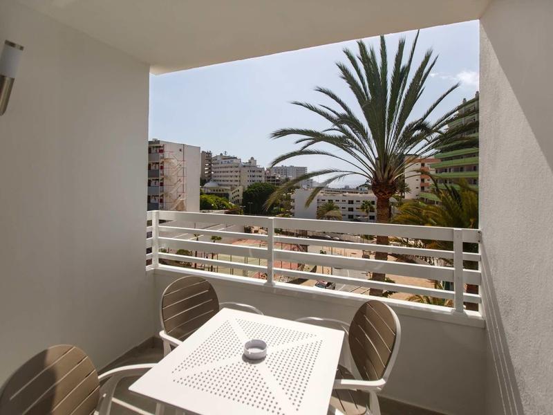 Balcon avec table et chaises donnant sur des palmiers et des bâtiments sous un ciel clair.