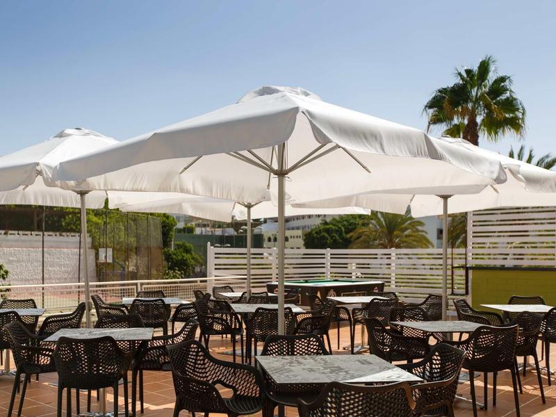 Restaurant en plein air avec parasols blancs et chaises noires, entouré de palmiers.