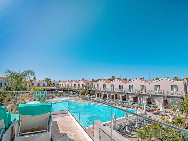 Nowoczesny basen hotelowy z leżakami i rzędami bungalowów pod niebieskim niebem.