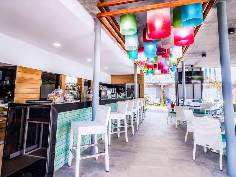 Nowoczesny bar hotelowy z kolorowymi lampami i białymi stołkami w jasnym wnętrzu.