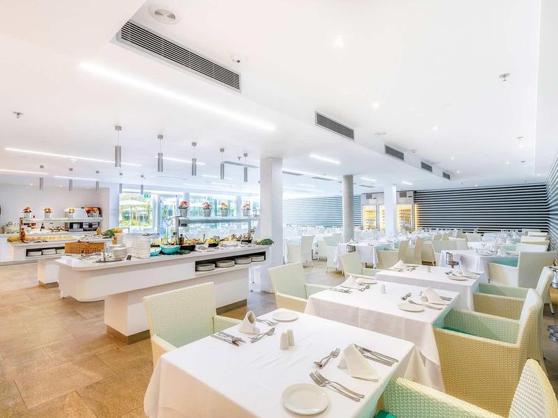 Nowoczesna restauracja z białymi nakrytymi stołami i jasnymi krzesłami w jasnym, przewiewnym pomieszczeniu.